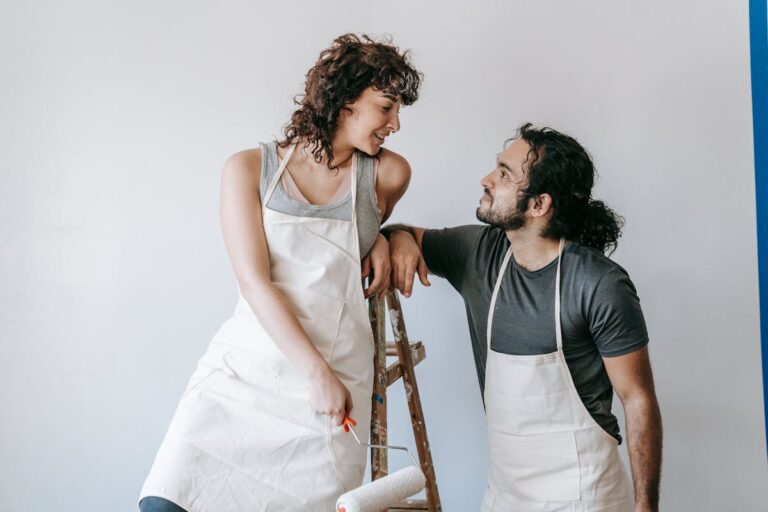 Smiling couple in aprons painting a wall with a roller, enjoying the renovation process.
